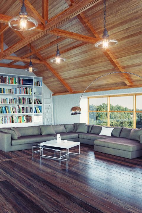 Mansarda luminosa con soffitto in legno, libreria bianca, divano grigio, tavolino centrale e lampade sospese, con vista panoramica attraverso ampie finestre.