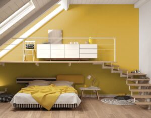Design innovativo di una camera da letto situata in un sottotetto. Il giallo senape domina la scena, con un letto moderno e dettagli come la sedia a pois, le scale di legno e i cassetti bianchi. La finestra in alto illumina l'intero ambiente, enfatizzando l'effetto accogliente e contemporaneo.