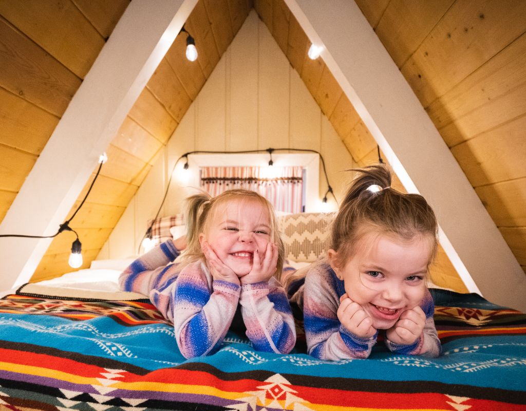 Due bambine sorridenti e spensierate si godono un momento di relax in una accogliente mansarda in legno, illuminata da luci soffuse. La vivacità dei loro sorrisi si riflette perfettamente nell'atmosfera calda e familiare dell'ambiente.