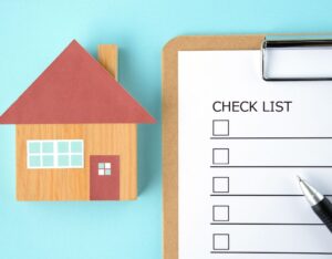 Un modellino di casa in legno posizionato su uno sfondo azzurro accanto a una clipboard che riporta una "CHECK LIST". Sulla lista sono presenti alcune caselle di controllo vuote. Una penna nera è appoggiata sulla clipboard, pronta per essere utilizzata.

