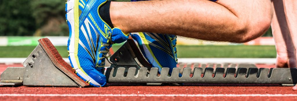 Atleta in scarpe da corsa blu e gialle posizionato sui blocchi di partenza su una pista d'atletica, pronto a correre